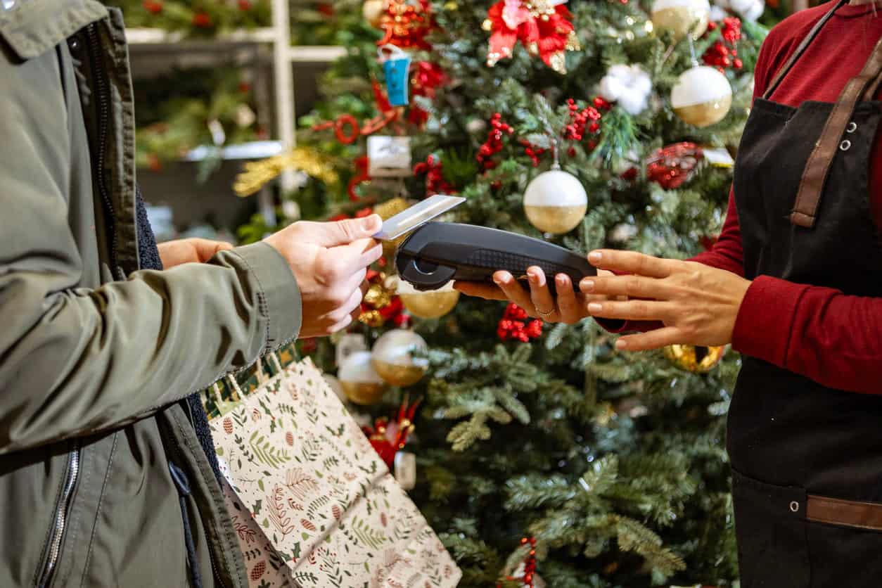 Shopper using a credit card payment terminal while holiday shopping in front of a decorated Christmas tree.