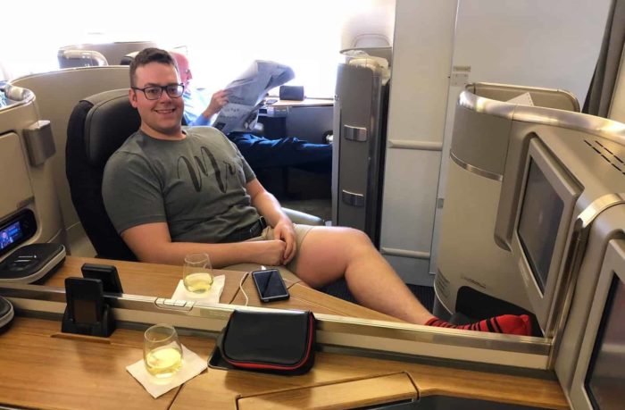 A man wearing glasses and a gray T-shirt relaxes in a spacious first-class airplane seat, enjoying his drink while traveling. Another passenger in the background reads a newspaper. The modern cabin features personal screens and stylish decor.
