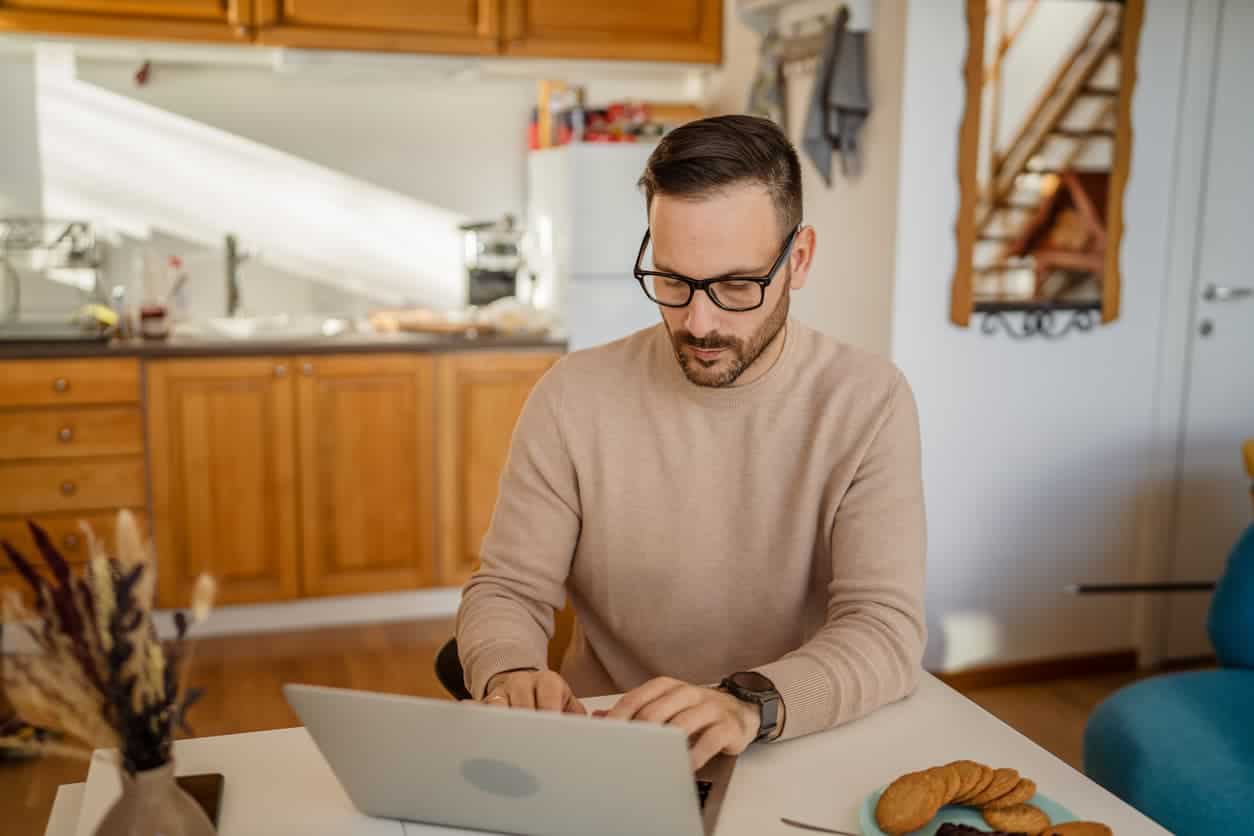 Man sitting at a kitchen table using a laptop.