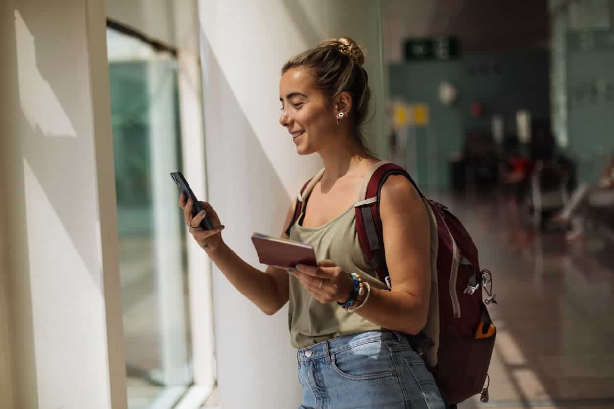 Woman at an airport holding a passport and phone, smiling while looking at her screen with a backpack on.