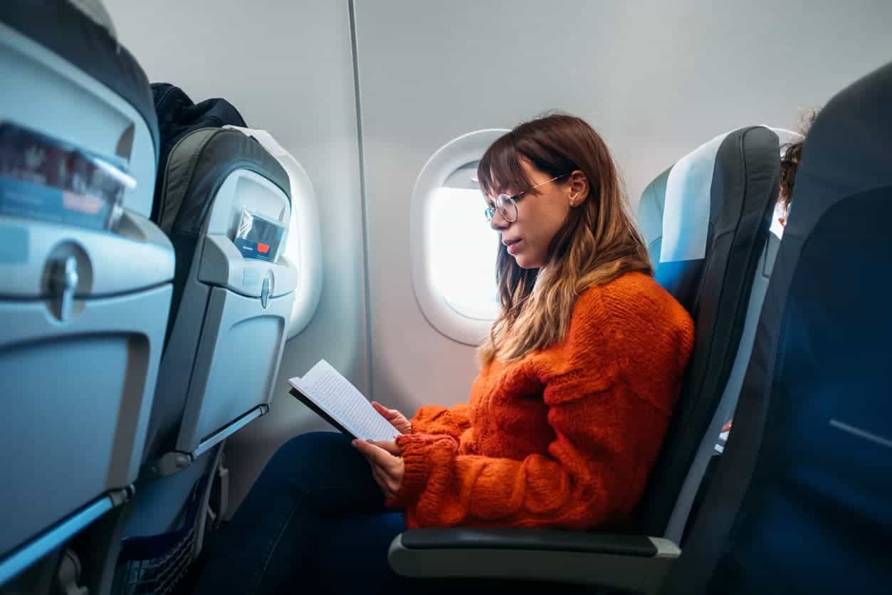 Woman sitting by an airplane window reading a book during a flight, wearing an orange sweater.