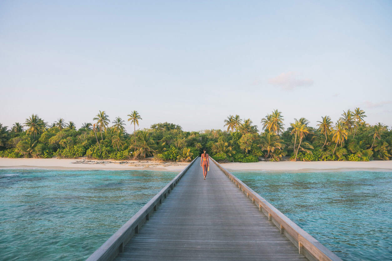 Woman walking along a wooden boardwalk toward a tropical island beach with palm trees and turquoise water in the Maldives