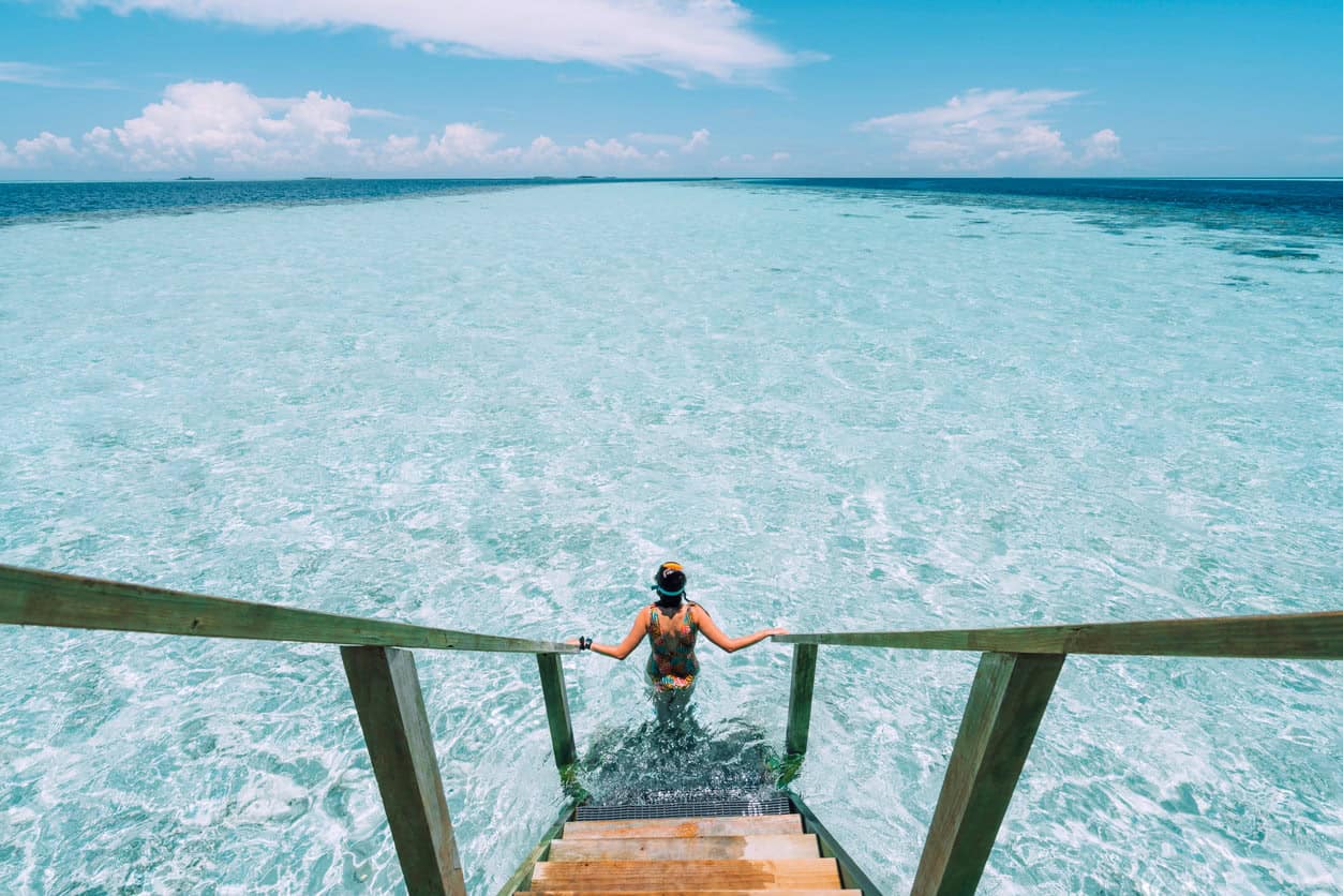 Woman entering crystal clear lagoon from wooden steps in the Maldives with shallow turquoise water and tropical horizon view
