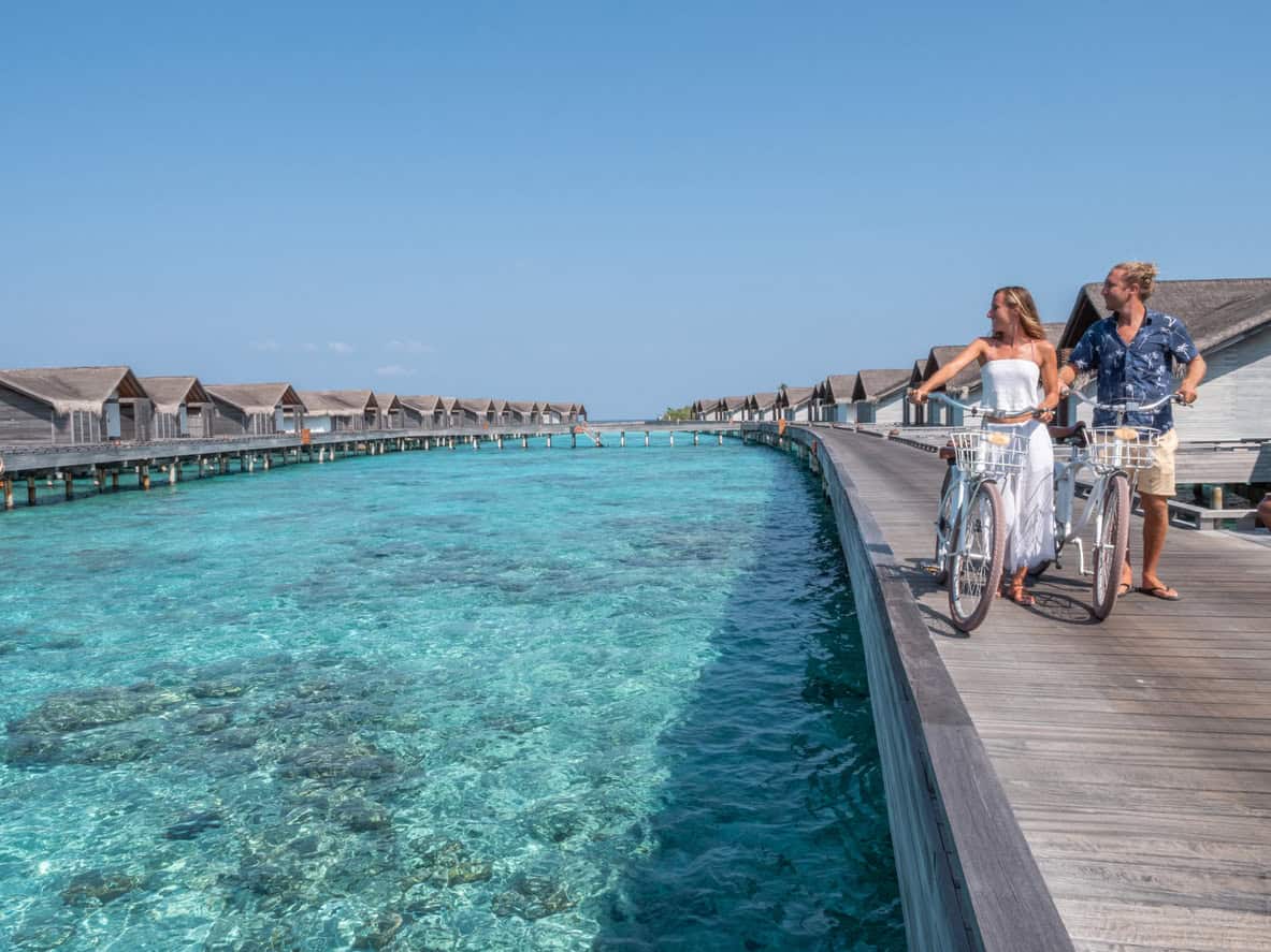 Couple riding bicycles along a wooden boardwalk with overwater villas in the Maldives and clear turquoise lagoon views