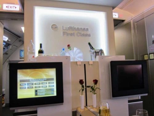 Lufthansa First Class cabin with illuminated sign, two screens displaying entertainment options, fresh roses in white vases, wine bottles, glasses, and elegant decor creates a luxurious in-flight atmosphere for those using miles or points.