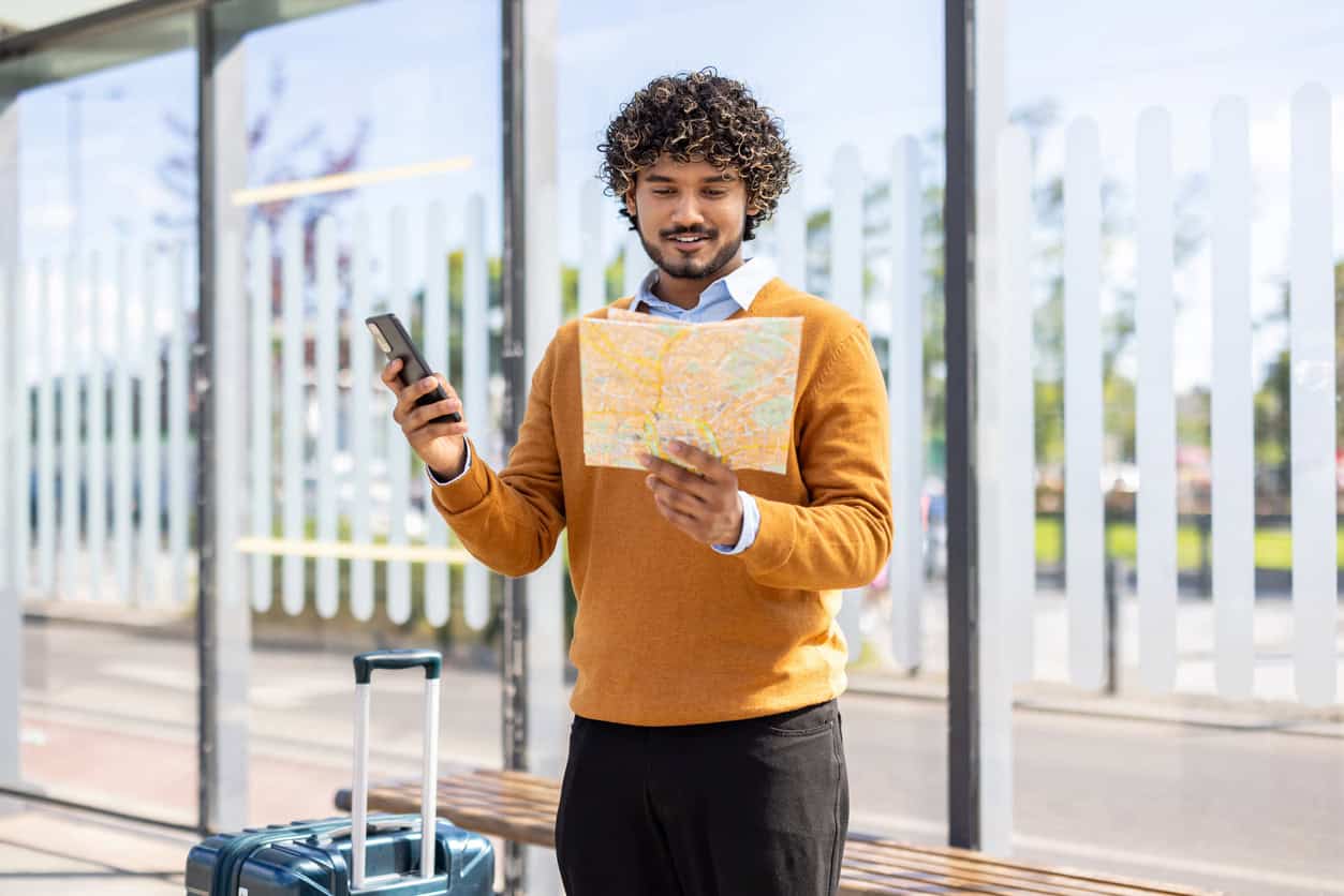 Traveler holding a map and smartphone while standing with luggage at a transit stop