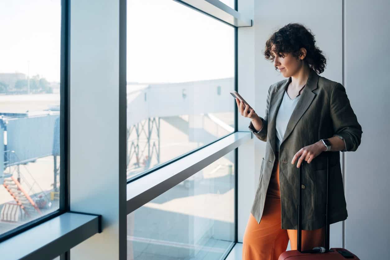Woman standing by a large airport window, checking her phone while holding the handle of a carry-on suitcase near a boarding bridge