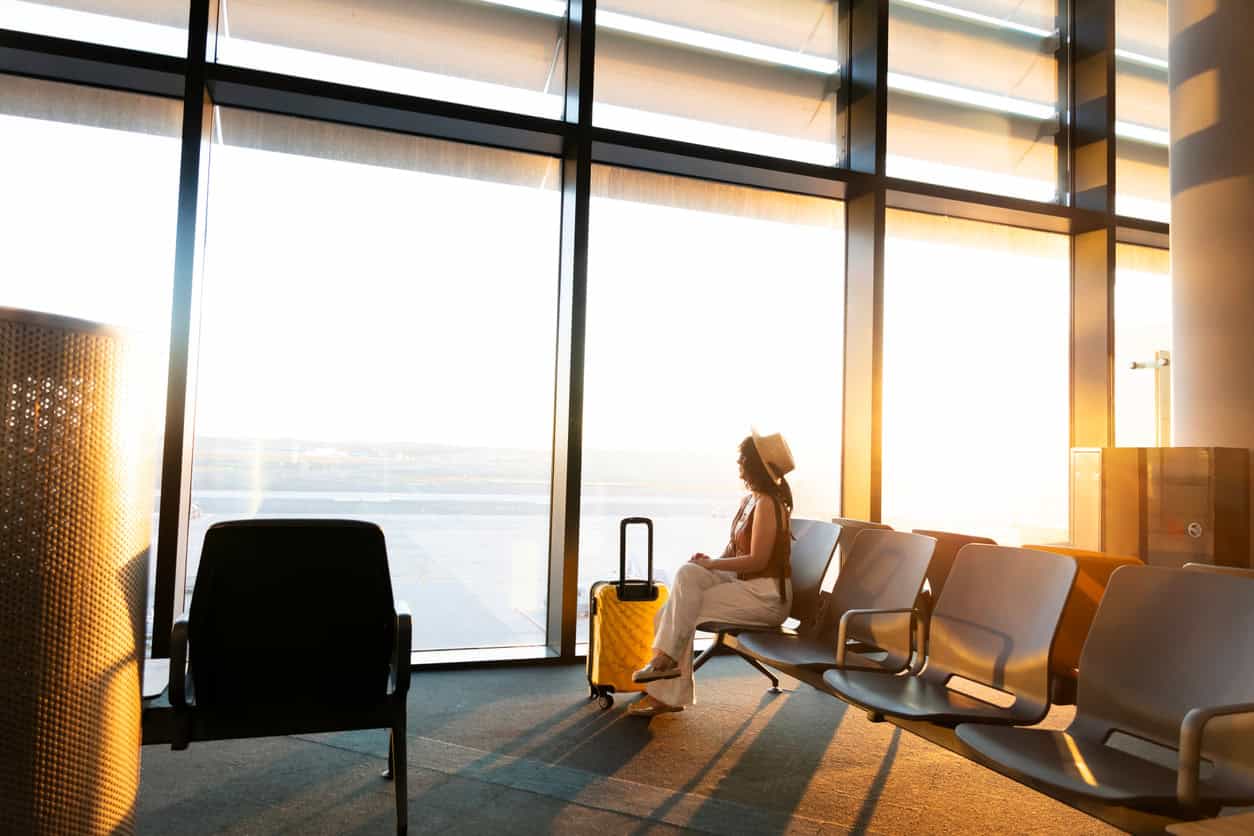 Woman sitting in an airport terminal at sunset with a yellow carry-on suitcase, waiting near large windows and empty seats