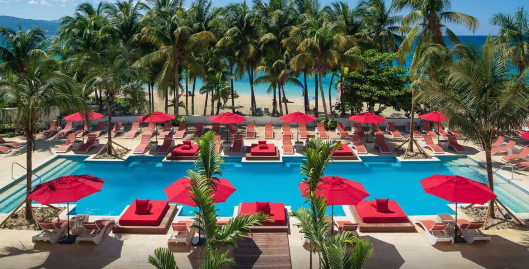 Tropical beachfront resort pool with red umbrellas, lounge chairs, and palm trees overlooking the ocean