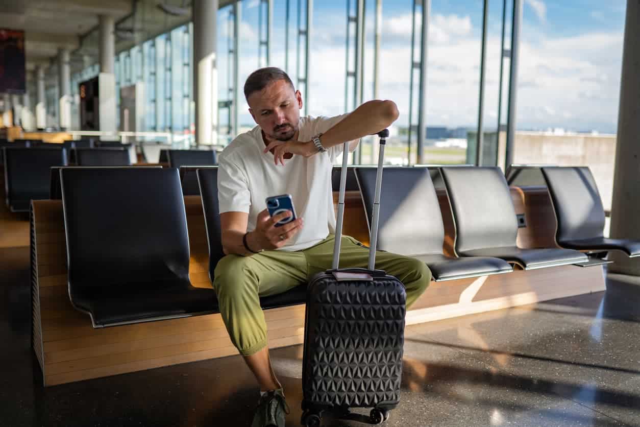 Frustrated traveler sitting at airport with luggage checking phone for flight updates