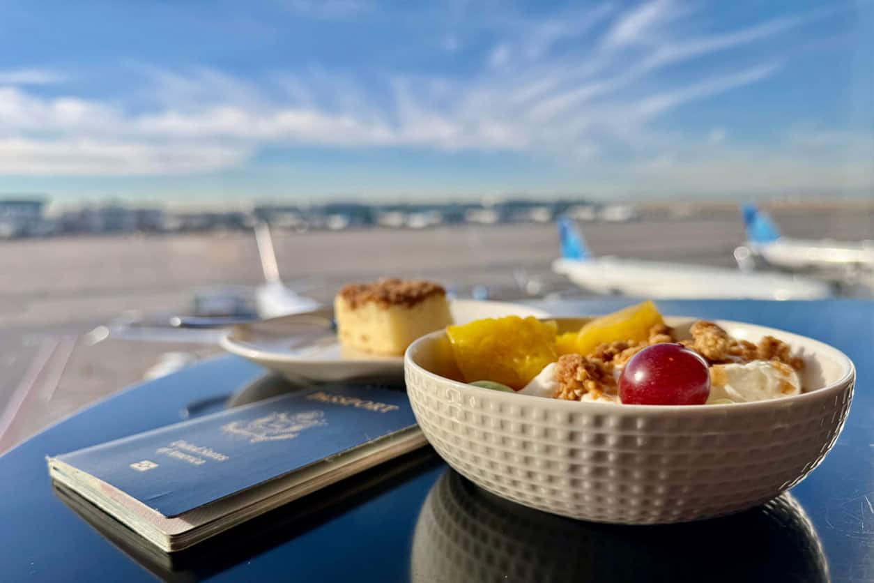 Breakfast with yogurt, fruit, and granola beside a passport on an airport lounge table overlooking airplanes on the runway