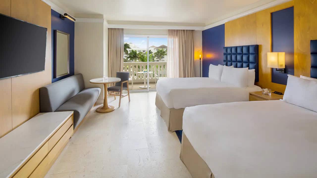 Bright ocean-view guest room at Hyatt Zilara Rose Hall in Jamaica with two double beds, modern blue-and-wood décor, and a private balcony overlooking tropical greenery