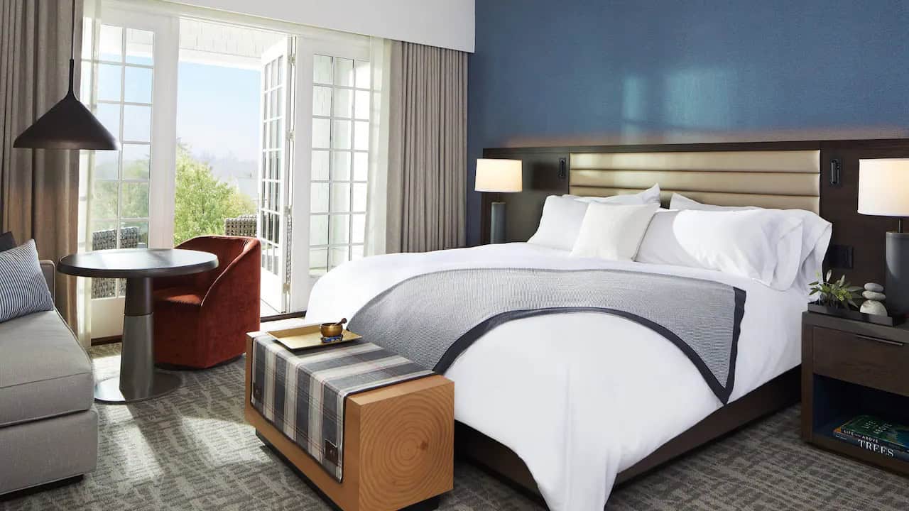 King guest room at Miraval Berkshires Resort & Spa featuring a plush bed, blue accent wall, modern sitting area, and French doors opening to a private outdoor terrace