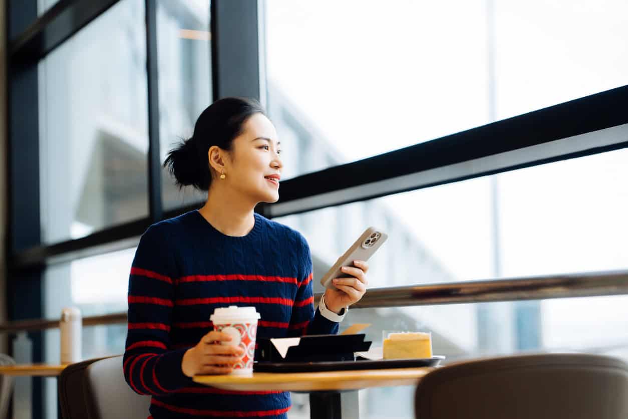 Woman sitting at an airport café table holding a smartphone and coffee with dessert while looking out the window
