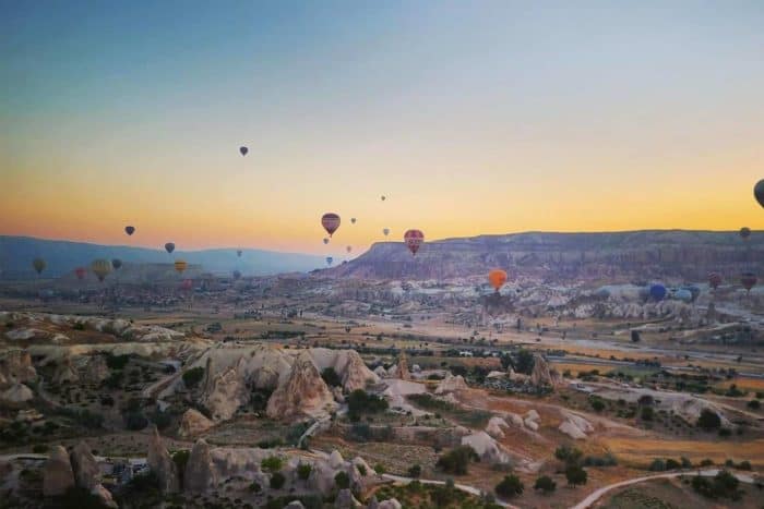 Dozens of colorful hot air balloons float above a rocky, scenic landscape at sunrise, perfect for travel enthusiasts collecting miles. The terrain is dotted with unique rock formations and sparse vegetation beneath a clear sky.