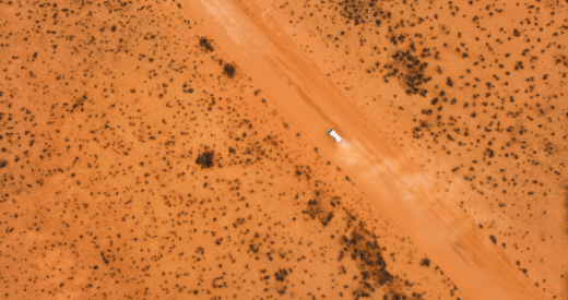 A white vehicle travels for miles along a straight, dusty road cutting through an arid, orange-brown desert landscape dotted with sparse shrubs and vegetation, seen from above.