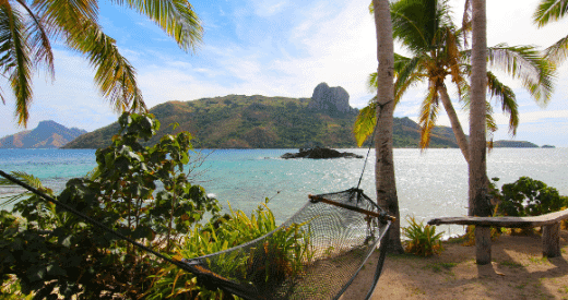 A hammock hangs between palm trees on a sandy beach, overlooking clear blue water and a lush, green island with hills in the background. Perfect for relaxing after miles of exploration or planning your next travel adventure.