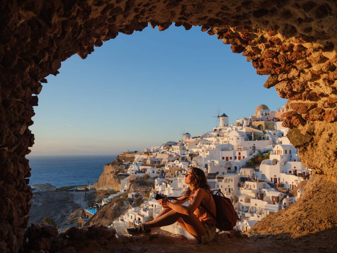 Traveler sitting inside a rocky cave overlooking white buildings and windmills of Oia Santorini at sunset with sea view
