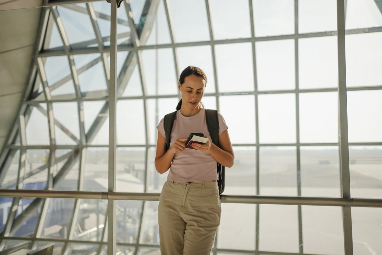 Female traveler at airport terminal looking at phone and boarding pass near large window with runway view