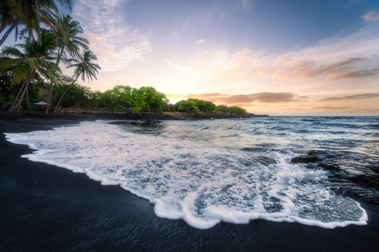Waves washing onto a black sand tropical beach at sunset with palm trees and a colorful sky