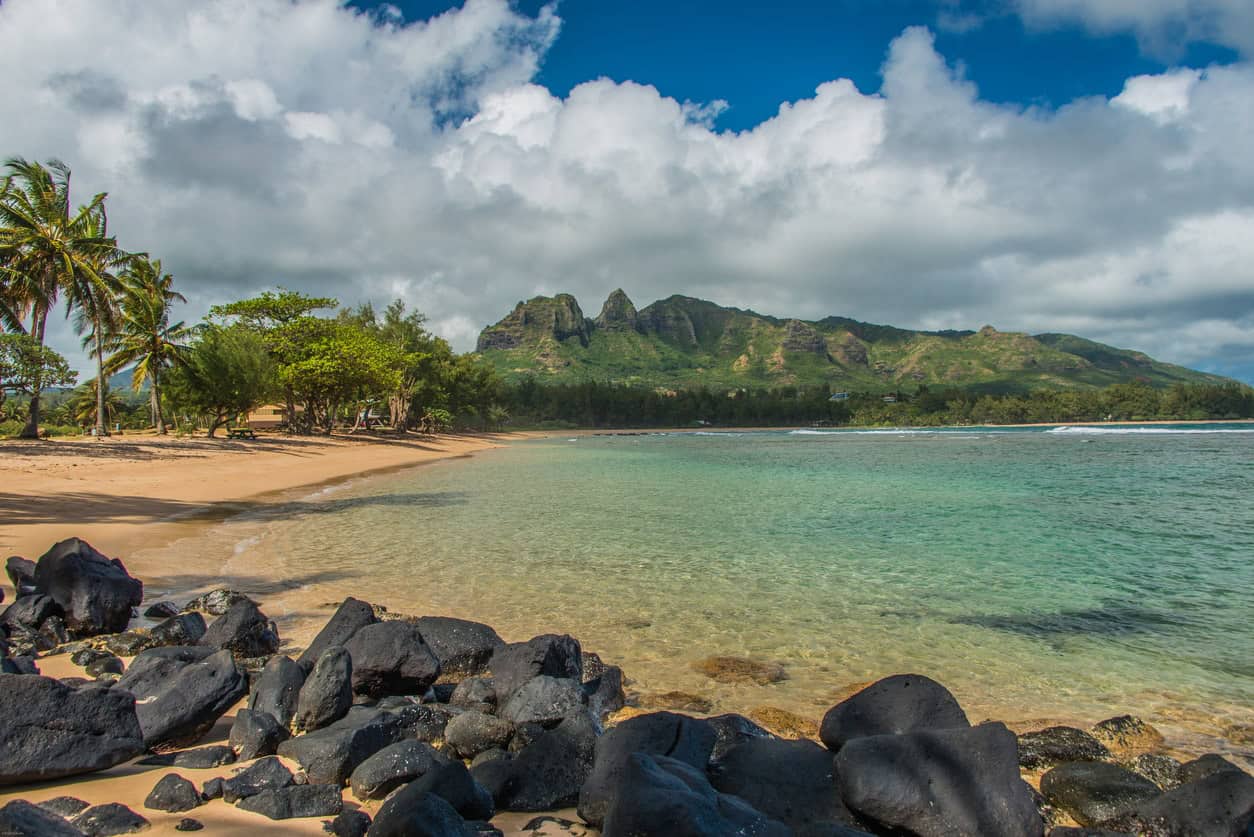 Clear tropical lagoon with rocky shoreline, sandy beach, palm trees, and lush green mountains under a cloudy blue sky
