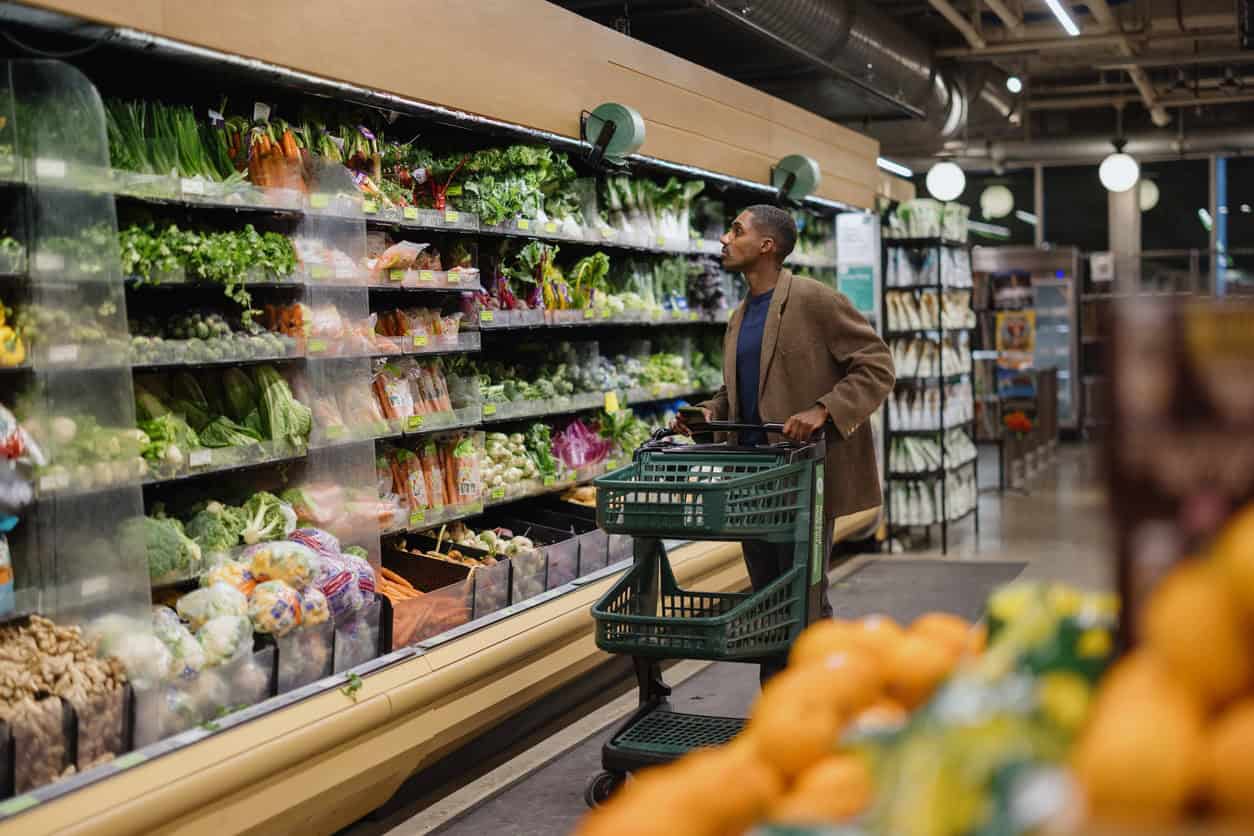 Man shopping in the produce aisle at a grocery store while pushing a green cart and selecting fresh vegetables.