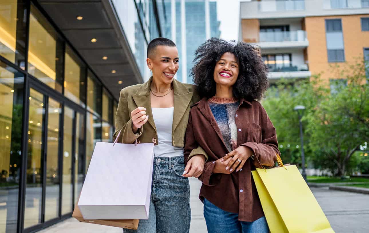 Two women walking outdoors with shopping bags, smiling and enjoying a day out together.