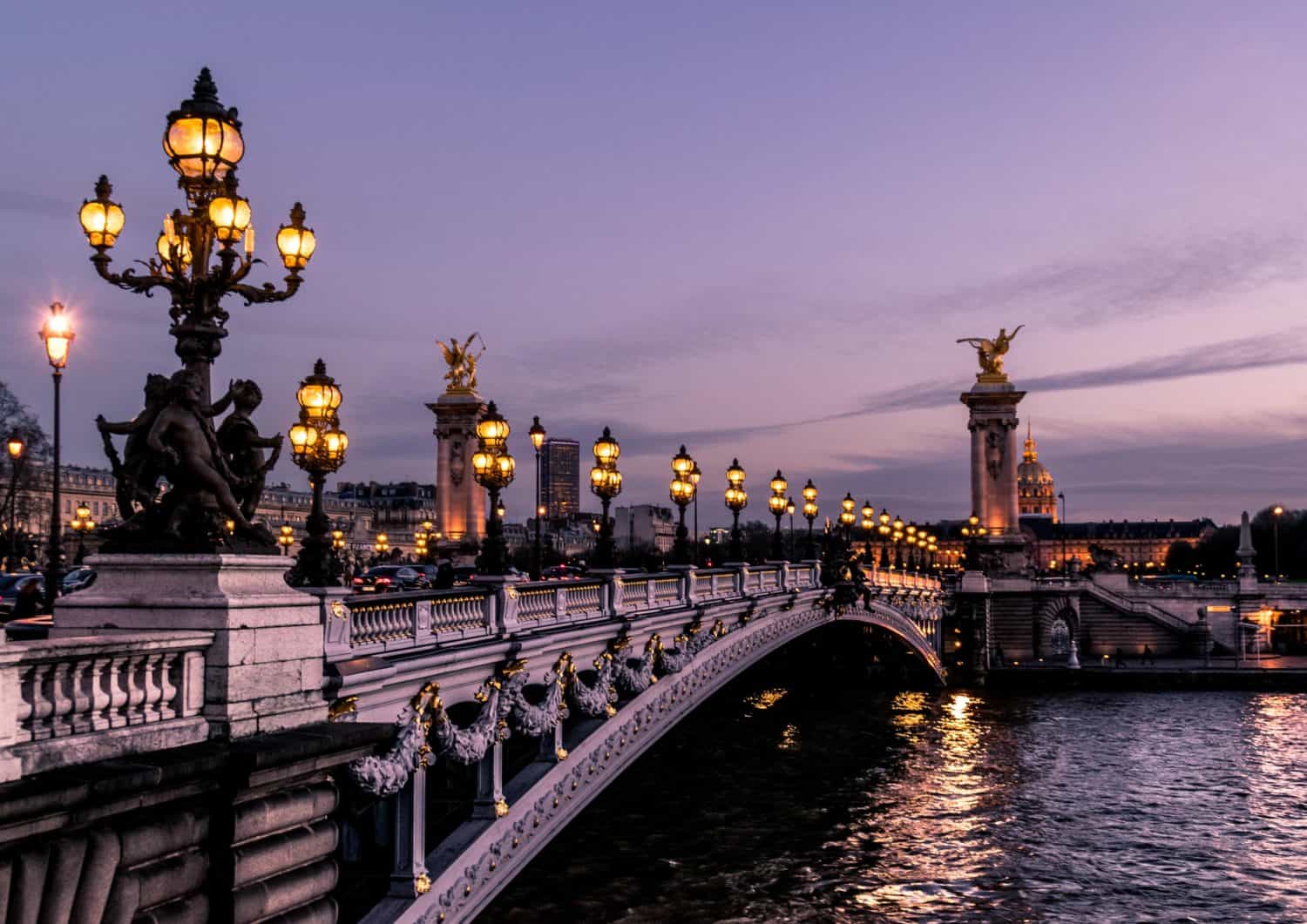 A view of the ornate Pont Alexandre III bridge in Paris at dusk, with golden street lamps glowing, decorative statues, and the river Seine flowing beneath. Perfect for travel enthusiasts seeking stunning city views as night falls.