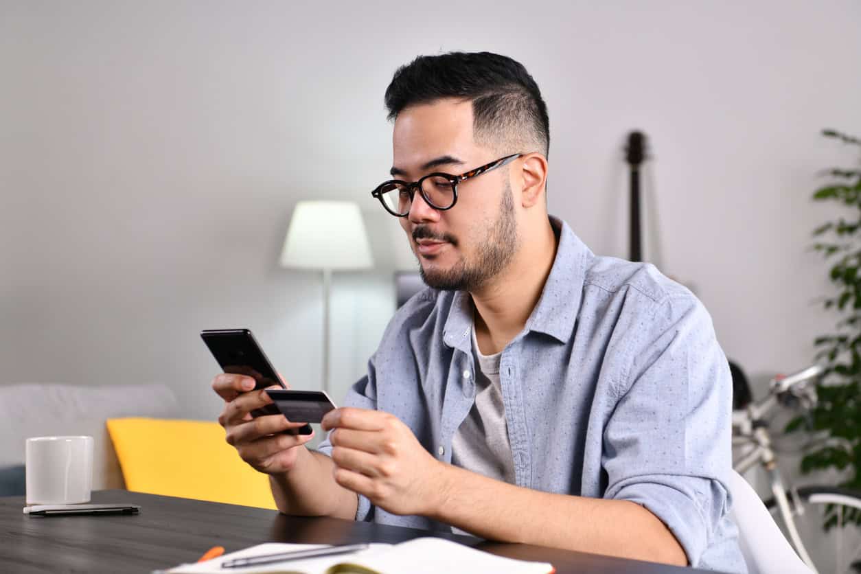 Man holding a credit card while using a smartphone at a desk at home