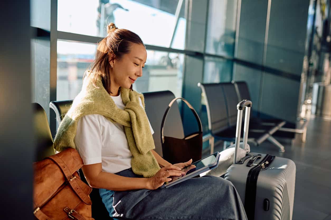 Traveler using a tablet to book a round-trip flight while waiting at an airport departure gate with luggage beside the seat.