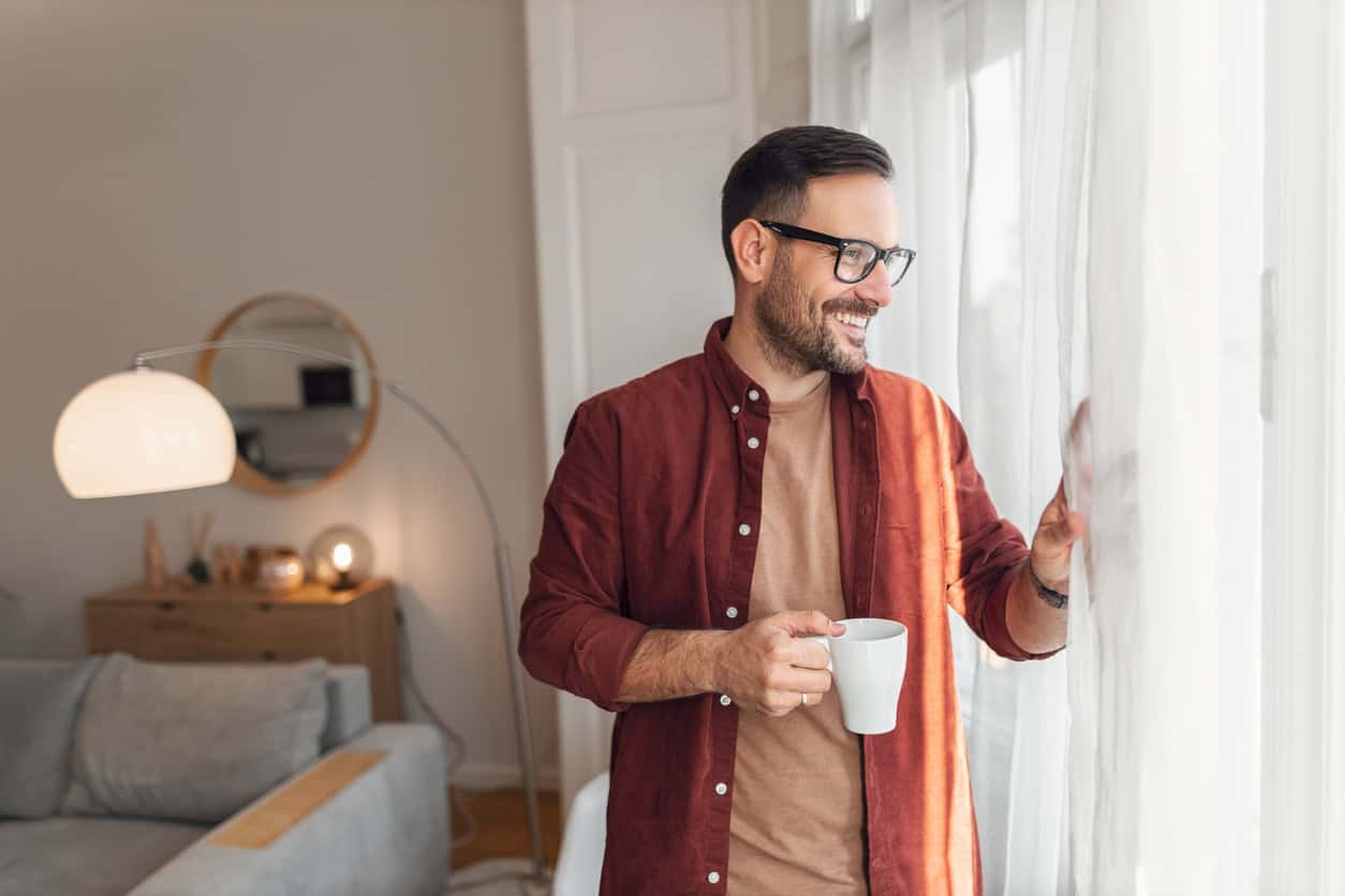 Smiling man holding a coffee mug while looking out the window in a cozy modern living room