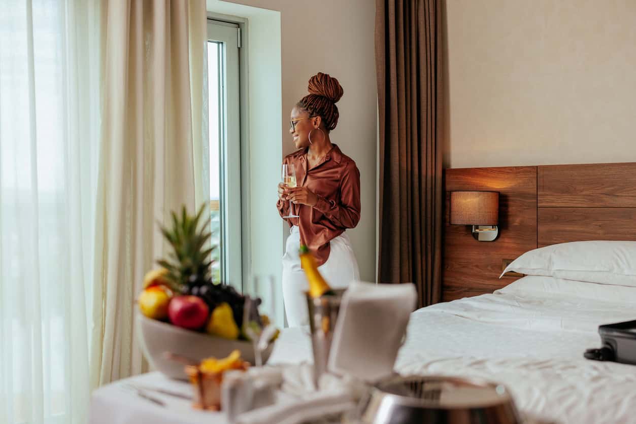 Woman standing by a hotel room window holding a glass of champagne with bed, fruit tray, and room service in the foreground