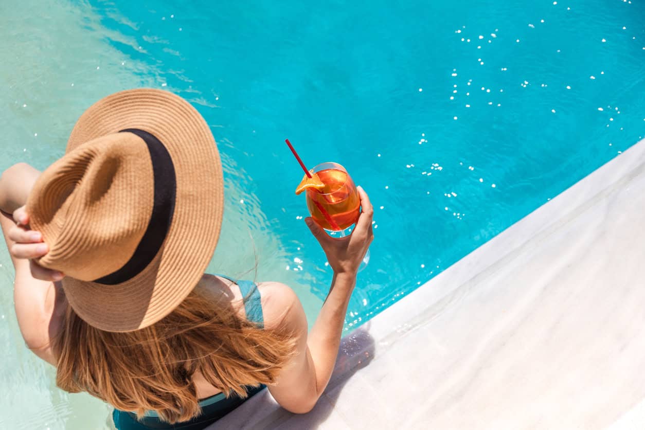 Woman in a hat sitting by a pool holding a cocktail with an orange slice garnish