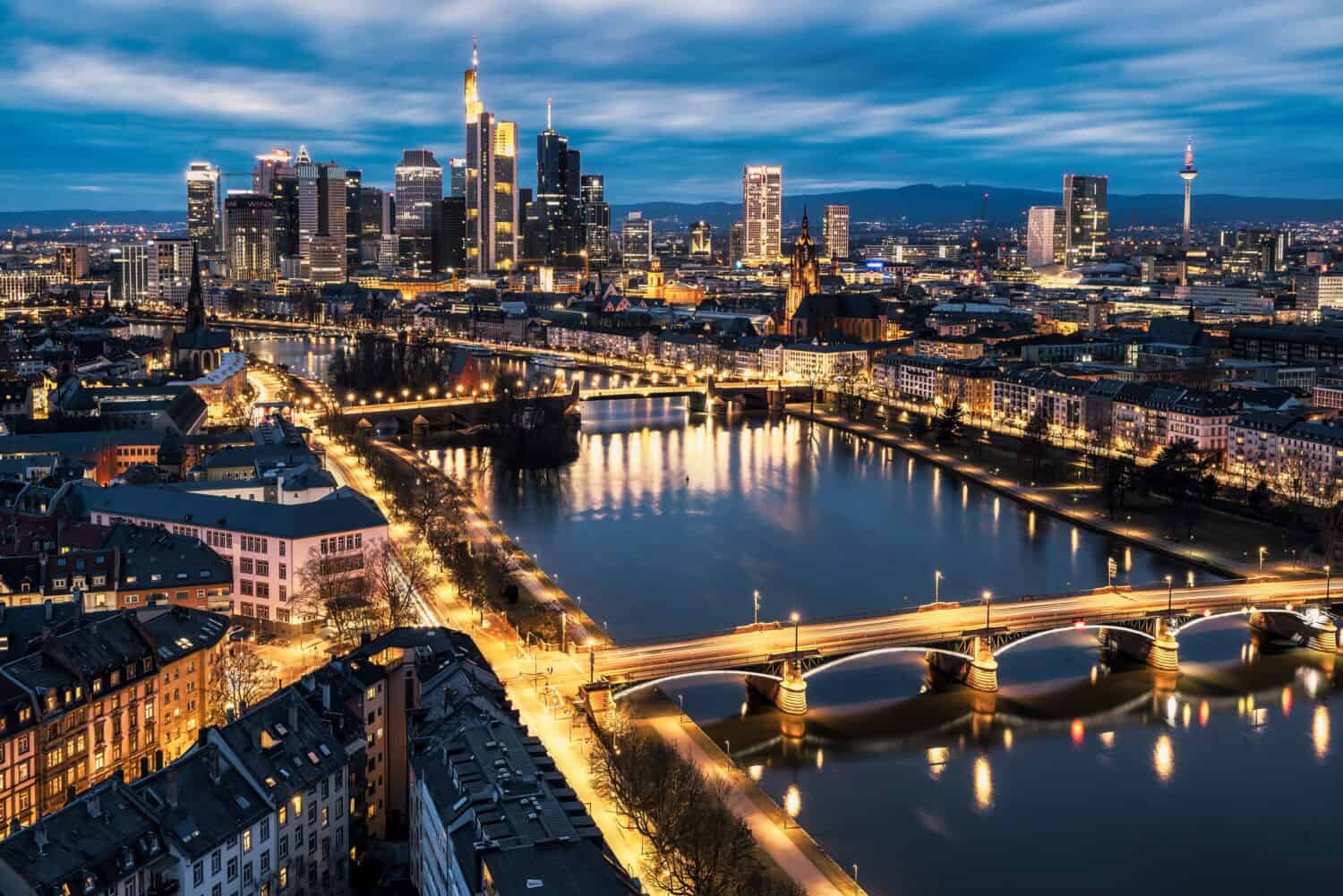 A cityscape of Frankfurt at dusk, showing illuminated skyscrapers, bridges crossing the Main River, and reflections of city lights on the water under a cloudy blue sky.