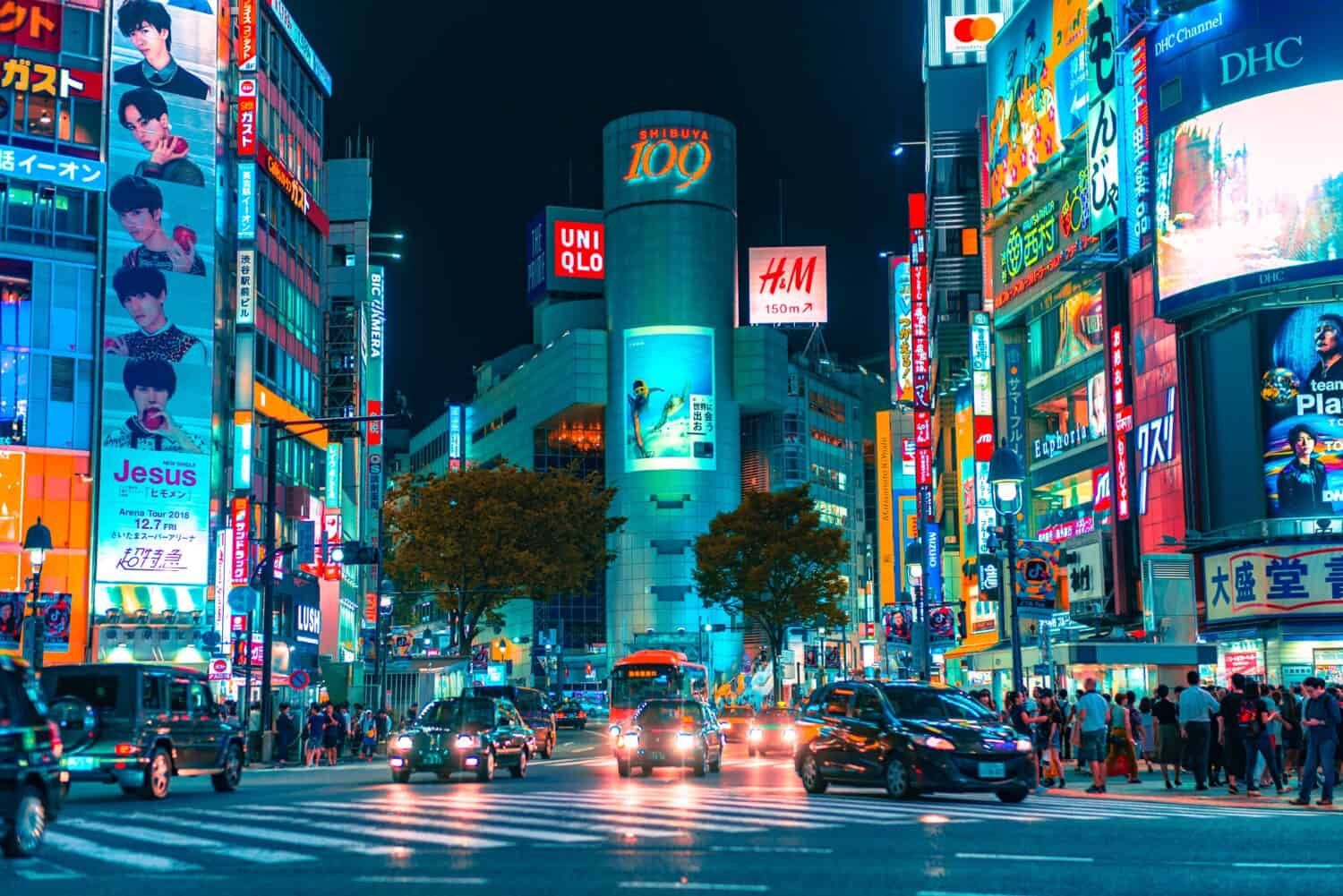 A vibrant city street at night in Shibuya, Tokyo, filled with colorful illuminated billboards, busy traffic, and crowds of people crossing the intersection.