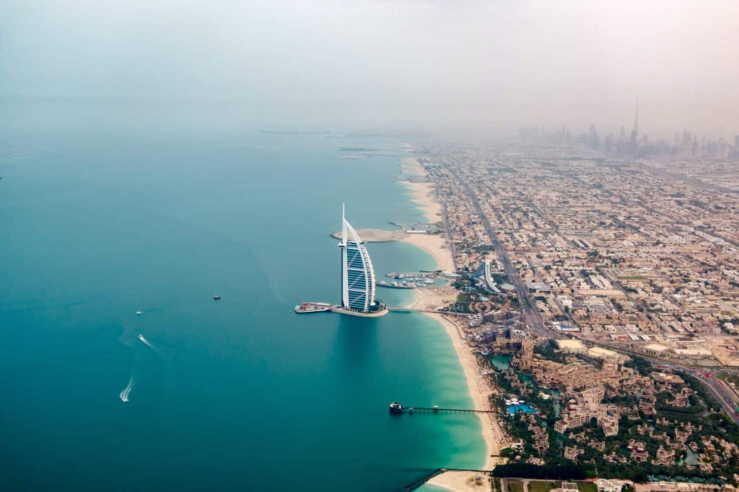 Aerial view of Dubai’s coastline featuring the Burj Al Arab hotel on a manmade island, turquoise waters, sandy beaches, and city buildings extending into the hazy distance.