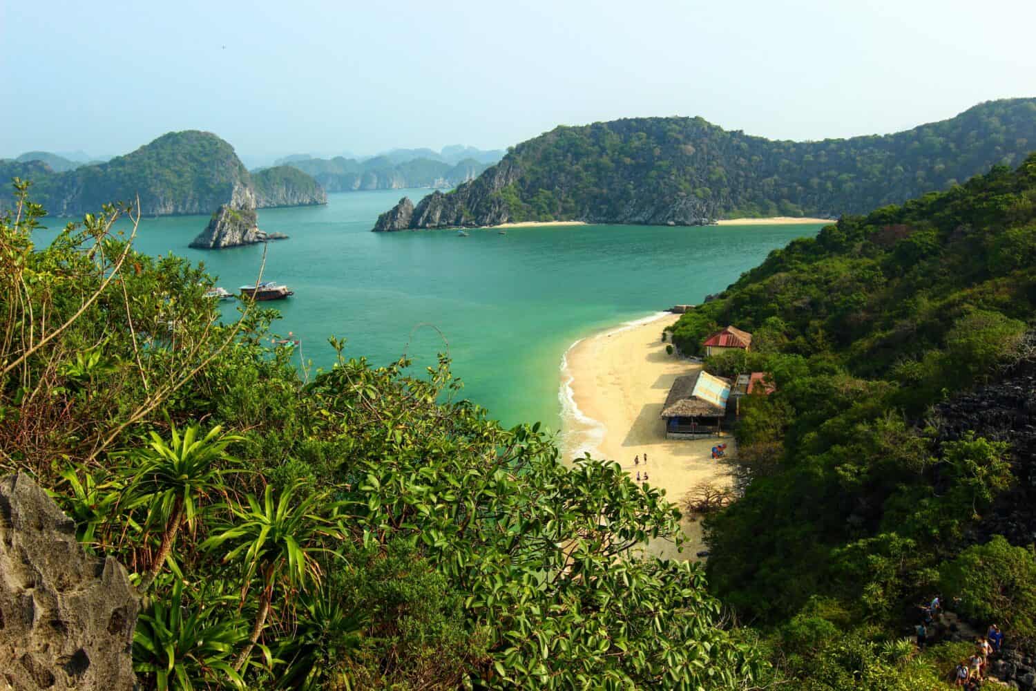 A scenic view of a tropical bay with clear blue-green water, sandy beaches, lush green hills, and small boats anchored near the shore, surrounded by rocky islands and dense vegetation.