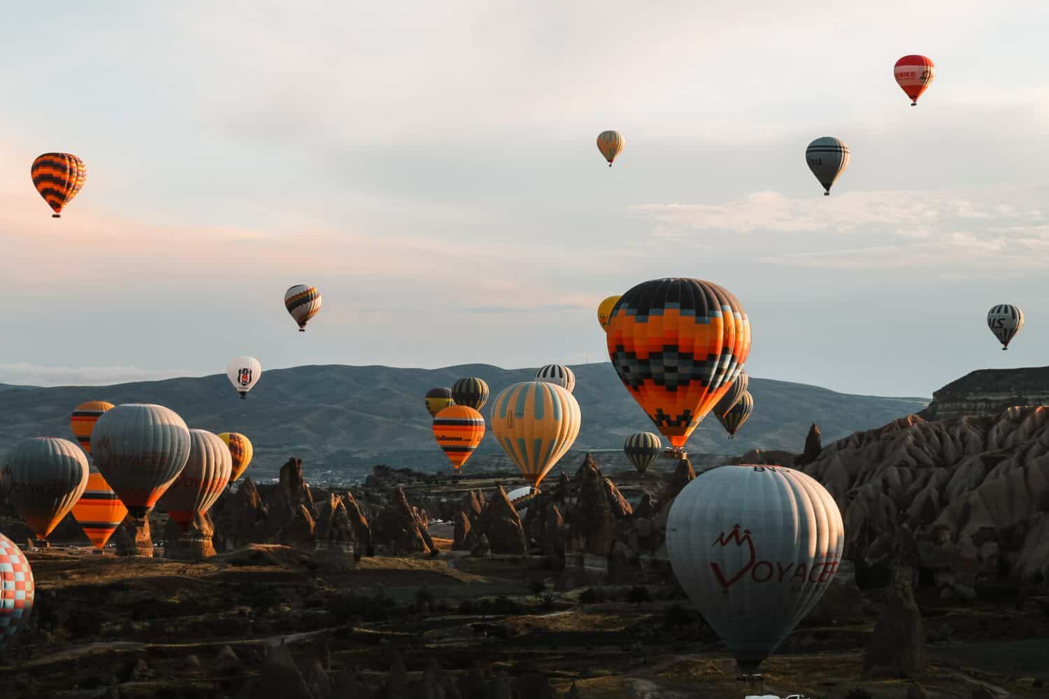 Hot air balloons of various colors and patterns float above a rocky landscape at sunrise, with distant hills and a partly cloudy sky in the background.