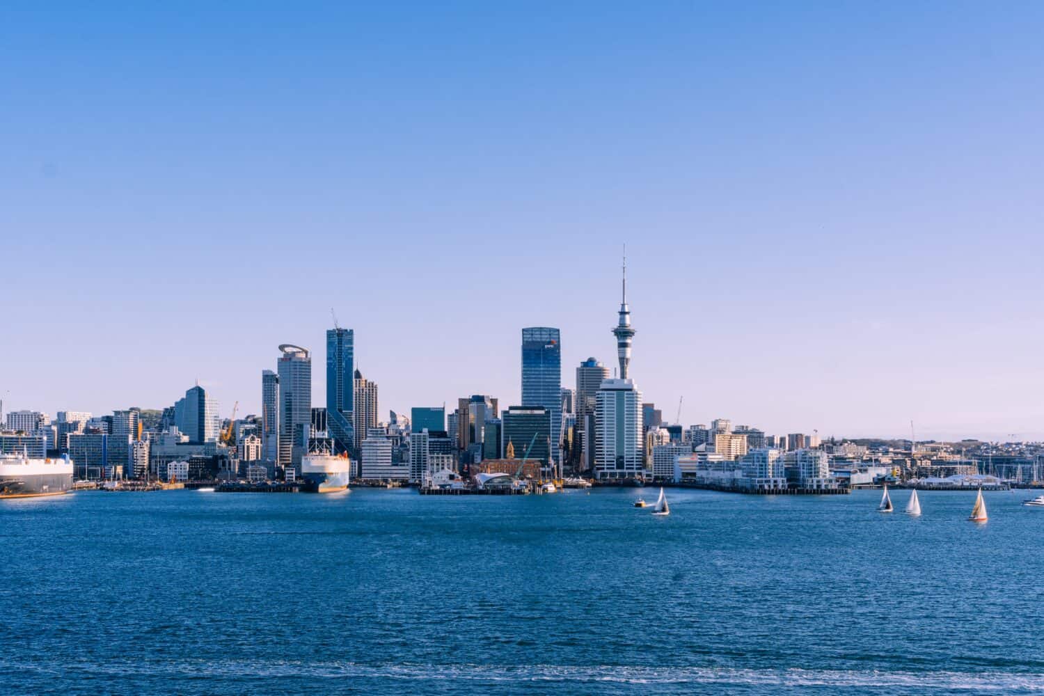 Auckland city skyline with modern high-rise buildings, the Sky Tower, and sailboats on blue harbor water under a clear sky.