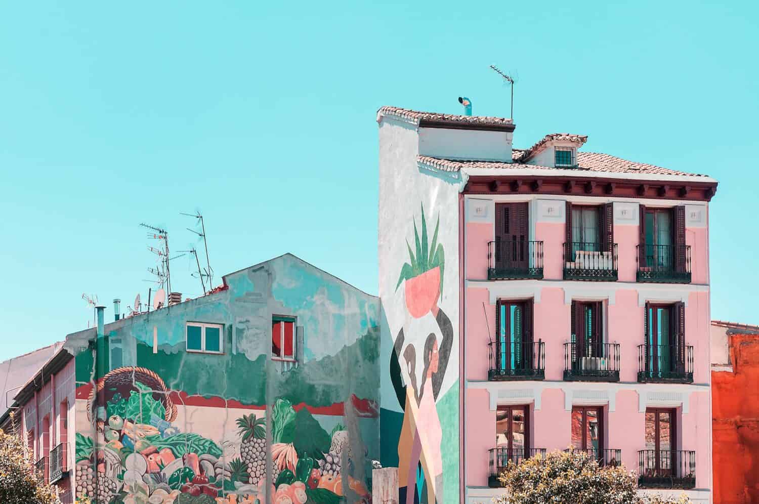 Colorful buildings with vibrant murals, including one with a group of raised hands holding a plant and another depicting a lush landscape, under a clear blue sky.