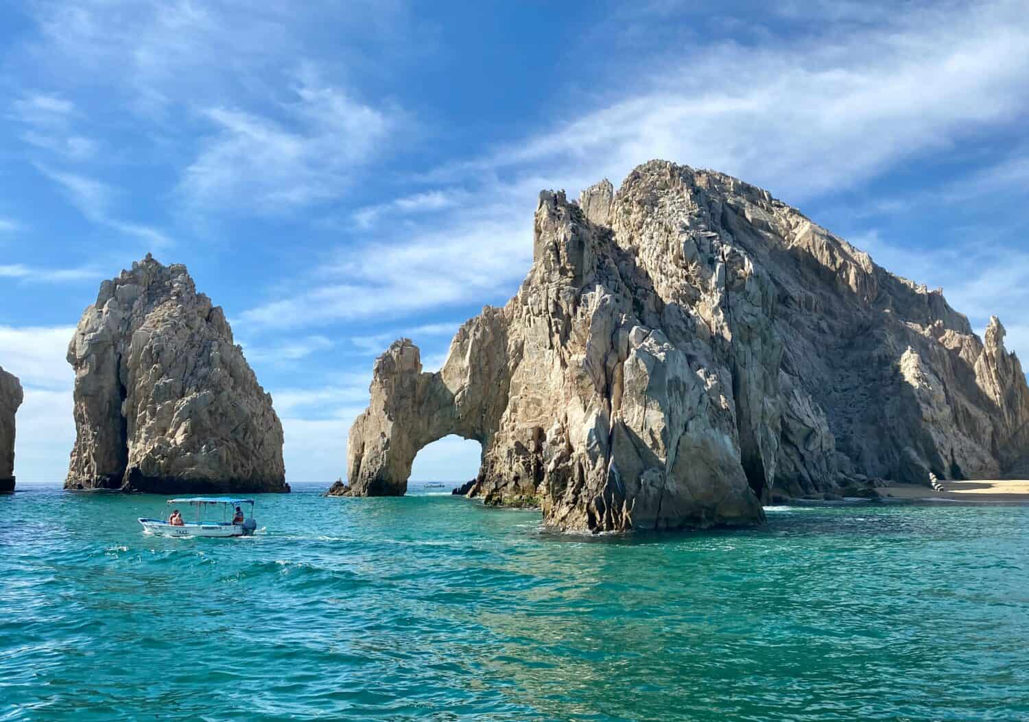 A small boat with people floats on clear turquoise water near a large rocky arch formation, with bright blue sky and some clouds above.