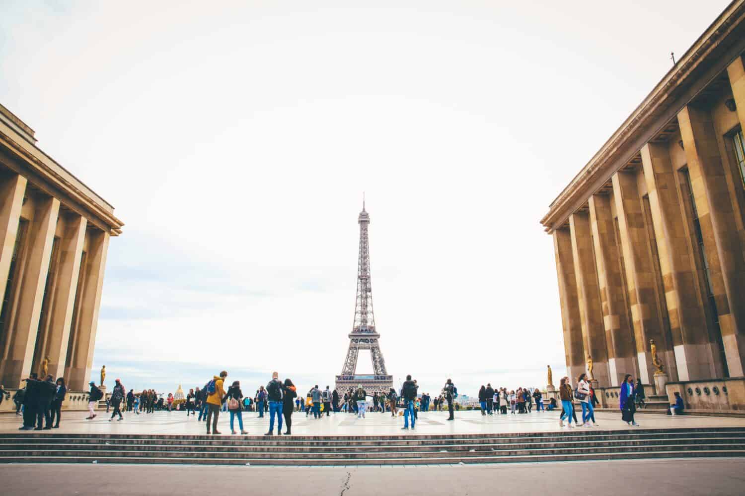 People gather on a large plaza with the Eiffel Tower in the background, framed by two tall classical buildings under a bright, cloudy sky in Paris.