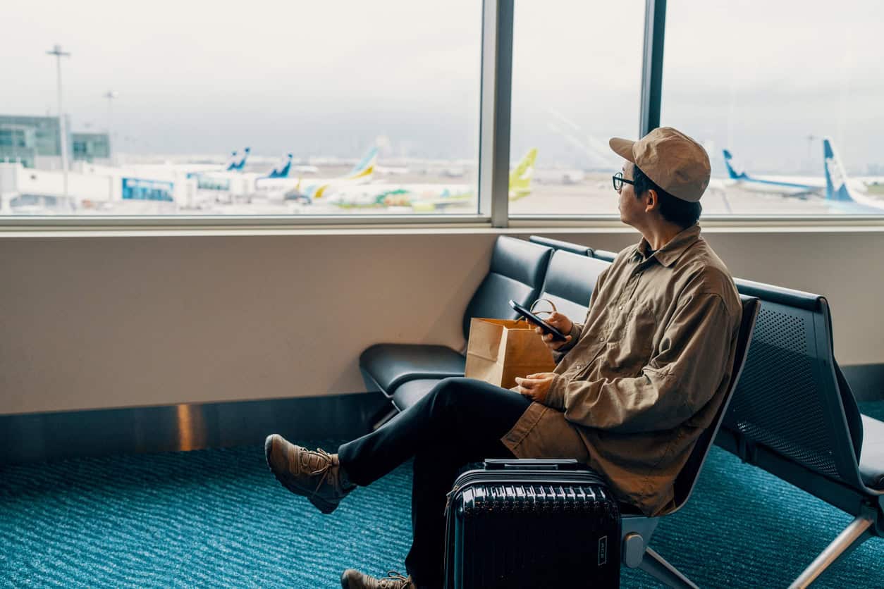 Traveler sitting in an airport terminal with luggage, looking out the window at airplanes while holding a smartphone