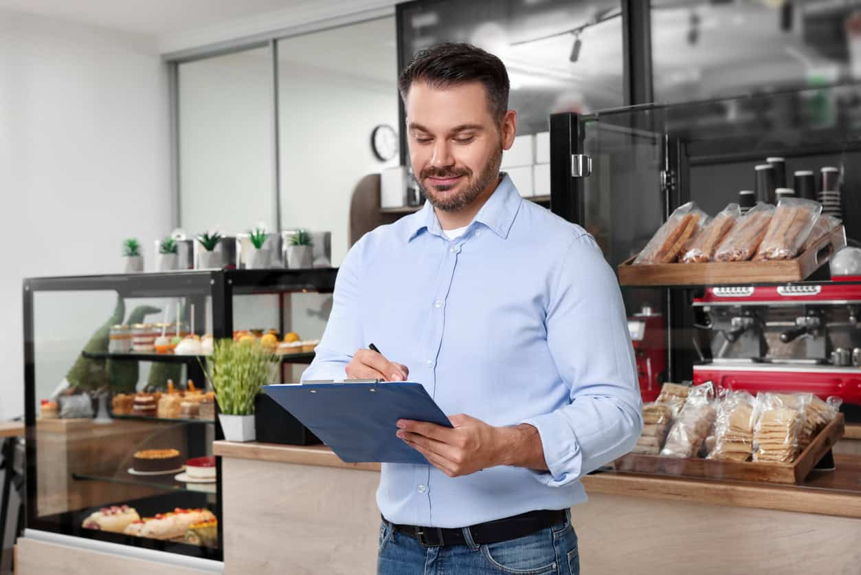 Small business owner checking inventory on a clipboard inside a café with baked goods and coffee equipment