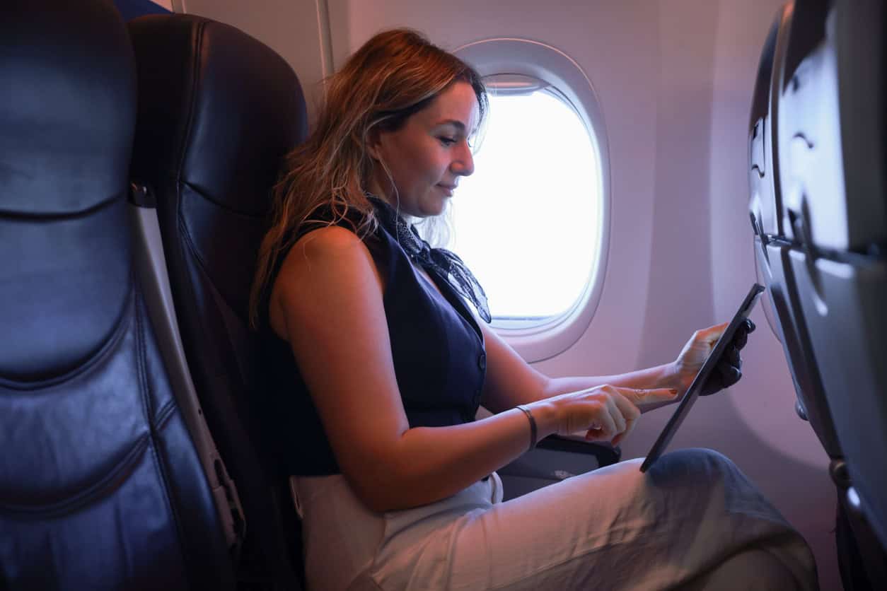 Female passenger sitting in an airplane seat using a tablet next to the window during a flight