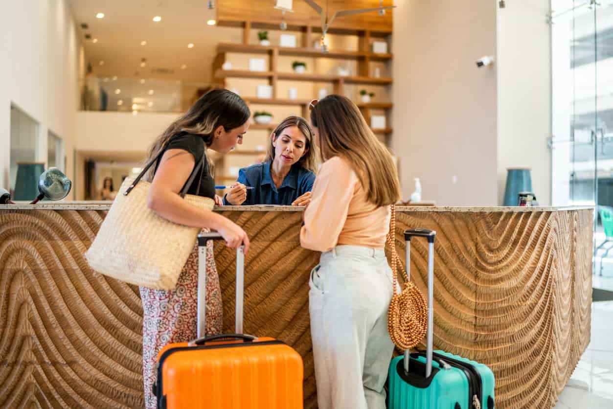 Two travelers checking in at a hotel front desk with suitcases.