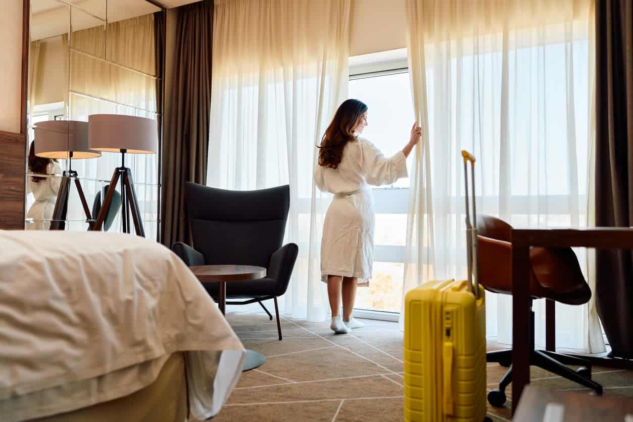 Woman in a hotel room opening the curtains with a suitcase beside her