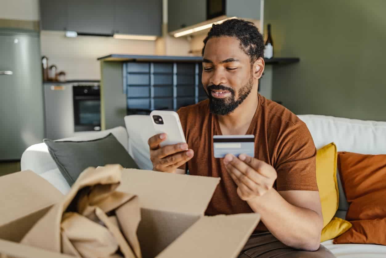 Man sitting at home using a credit card and smartphone to make an online purchase with an open package nearby.