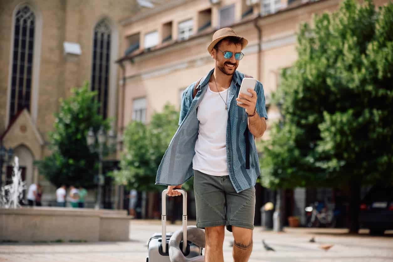 Man walking through a city street in summer attire while pulling a suitcase and checking his phone.