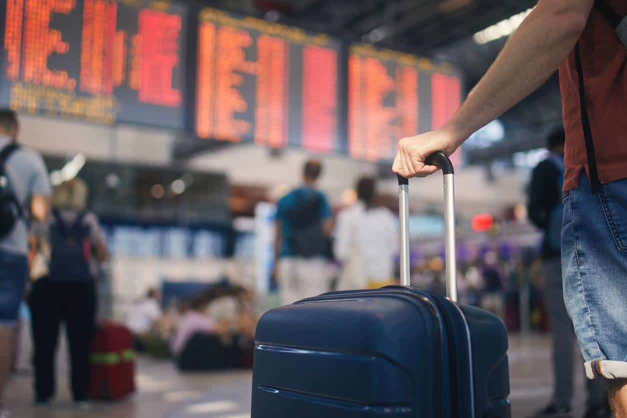 Traveler holding a suitcase in front of a flight departure board at a busy airport terminal